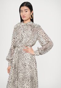 Hosszú ujjú ruha áttetsző anyagból, krémalapú barna pöttyös mintával. Gallérral, gombos elülső résszel és ráncolt ujjal rendelkezik.