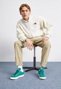 Jacket de fleece branco com fecho éclair e um logotipo preto, calças bege, e ténis verdes com solas brancas e marcação. Textura suave e ajuste relaxado.