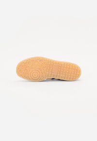 Gumová podrážka tenisky ve světle hnědé barvě, s texturovanými vzory protiskluzu a kruhovými akcenty. Modrý svršek viditelný v profilu.