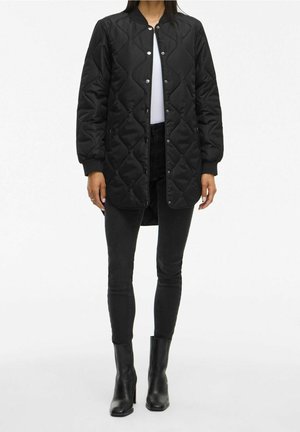 Person trägt eine schwarze gesteppte lange Jacke über einem weißen Top, schwarze enge Jeans und schwarze Stiefeletten mit Absatz und steht auf weißem Hintergrund.