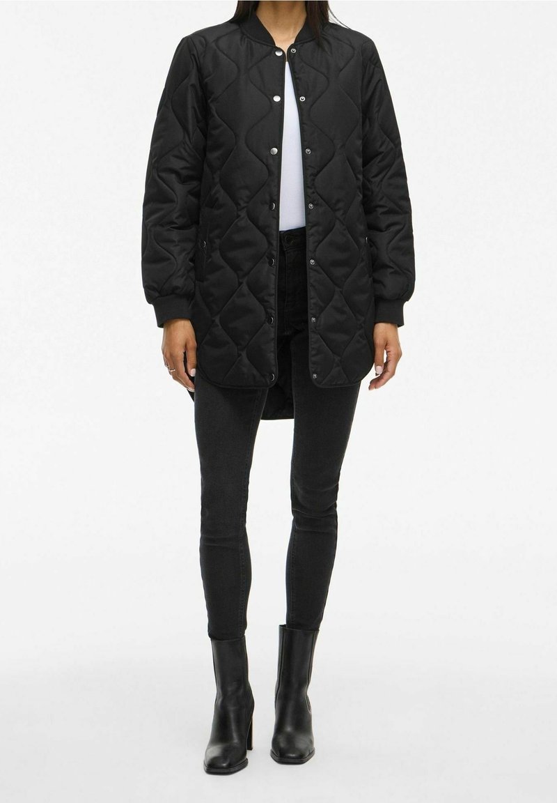 Personne portant une longue veste matelassée noire par-dessus un haut blanc, un jean skinny noir et des bottines noires à talons, debout sur un fond blanc.