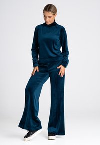 Turkusowy aksamitny dres z wysokim kołnierzem i długim rękawem oraz spodniami typu wide-leg. Miękka tekstura, czarne sneakersy uzupełniające strój.