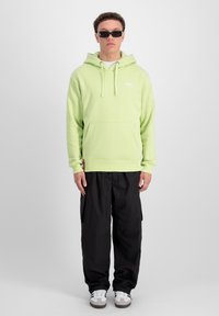 Felpa verde chiaro con tasca frontale, cappuccio con cordino e logo. Abbinata a pantaloni cargo neri e sneakers grigie e bianche.