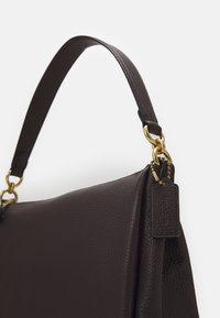 Sac à main en cuir marron foncé avec une sangle courte, des ferrures dorées et une étiquette en cuir assortie fixée par une petite chaîne à billes dorée.