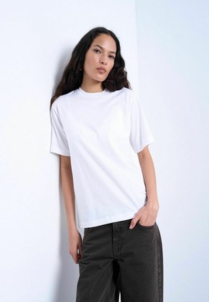 Woman with long dark hair leans against white wall, wearing a plain white T-shirt and black pants, one hand in pocket.