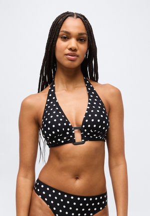 Femme aux longues tresses portant un haut de bikini noir à col licou et un bas de bikini assorti à pois sur un fond uni.