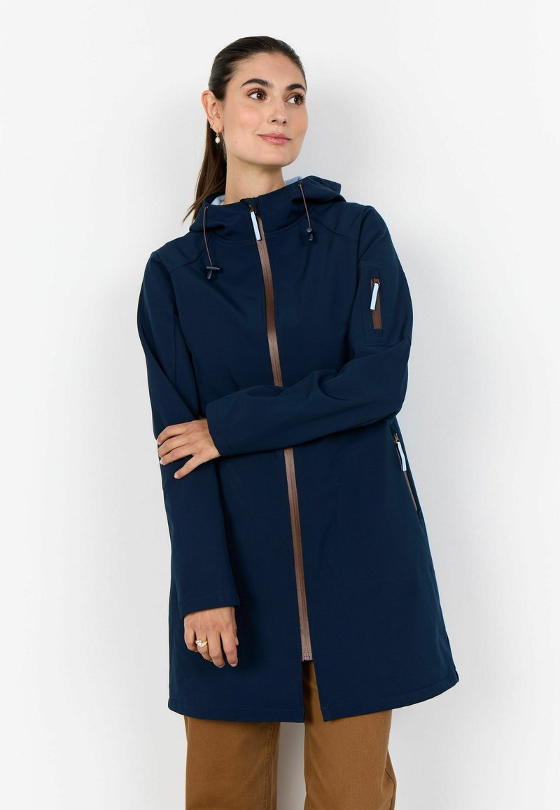 Žena v tmavomodrej bunde s kapucňou a hnedým zipsom, v béžových nohaviciach, stojaca s prekríženými rukami a mierne otočená na stranu.