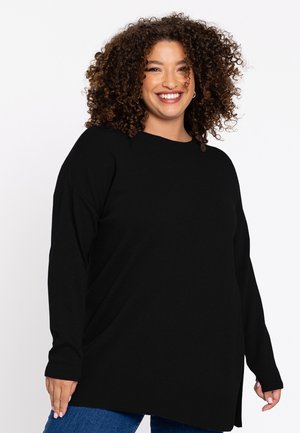 Smilende kvinde med krøllet hår iført en sort langærmet trøje og blå jeans stående foran en hvid baggrund.