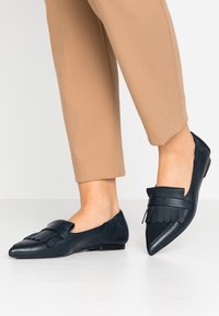 Mocassins en cuir bleu marine avec un bout pointu, dotés d'une frange décorative et d'un talon plat. Surface texturée et finition lisse sur l'ensemble.