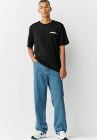 Camiseta de algodón negra con cuello redondo y un logo blanco, combinada con unos jeans de denim de corte relajado de color azul claro y zapatillas negras y blancas.