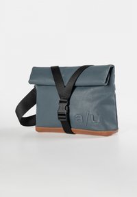 Gray waterproof bag with a fold-over top, black strap, and brown base. Features embossed brand initials and adjustable buckle.