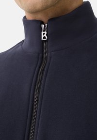 Mand iført en marineblå zip-up jakke med høj krave, lynlåstrækkeren med et metal bogstav "B" synligt nær halsen.