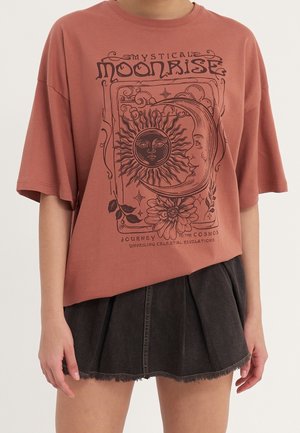 Persoon draagt een loszittend, roestkleurig t-shirt met een illustratie van zon, maan en bloem en een zwarte geplooide denim mini-rok.