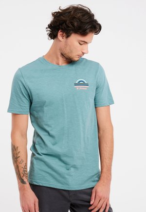 Jonge man met donker krullend haar, gekleed in een lichtblauw T-shirt met opdruk en donkere broek, kijkt omlaag waarbij een tatoeage op zijn linkeronderarm zichtbaar is.