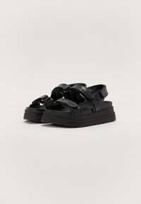 Sandalias negras con acabado brillante, que cuentan con correas anchas y cierres de Velcro. La suela de la plataforma es gruesa y tiene una superficie texturizada para mayor tracción.