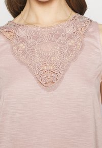 Ljust rosa utan ärmar topp med intrikata spetsdetaljer vid halsringningen, texturerat tyg och en något lös passform.
