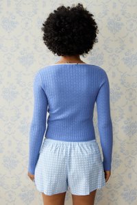 Figurformad ribbad tröja i ljusblått med långa ärmar, kombinerad med ljusblå rutiga shorts. I bakgrunden syns ett texturerat blommönster.