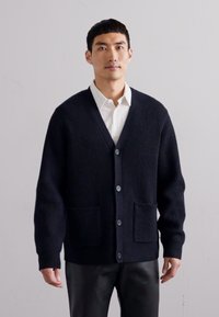 Tumesinine ribakootud kardigan, millel on V-kaelus, nööbist ees, kaks pealiskotti ja pikad varrukad. Pehme tekstuur ja vabalt istuv lõige.