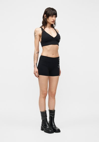 Femme debout de face portant un soutien-gorge de sport noir, un short noir ajusté avec du texte blanc, et des bottes noires mi-mollet sur un fond uni.