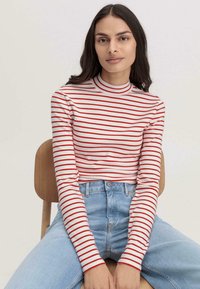 Langarmshirt mit rot-weißen horizontalen Streifen, hohem Kragen und weichem Stoff. Kombiniert mit hochgeschnittenen blauen Denim-Jeans.