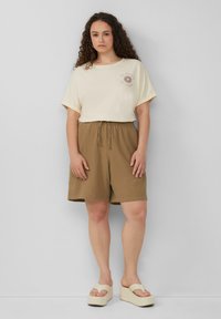 Lichtbeige katoenen T-shirt met opgerolde mouwen en zonnenafbeelding, gecombineerd met gestructureerde bruine shorts met een elastische tailleband. Beige platform sandalen.