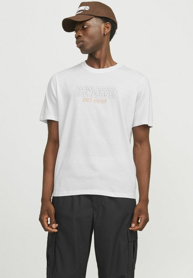 Hvid bomulds-T-shirt med teksten "Jack & Jones Ninety Nineteen" i grå med orange accent. Sammen med sorte cargo bukser.