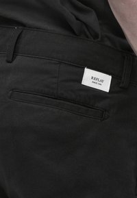 Μαύρο παντελόνι με πίσω τσέπη και λευκή ετικέτα μάρκας που γράφει "REPLAY SINCE 1981" κοντά στη μέση.