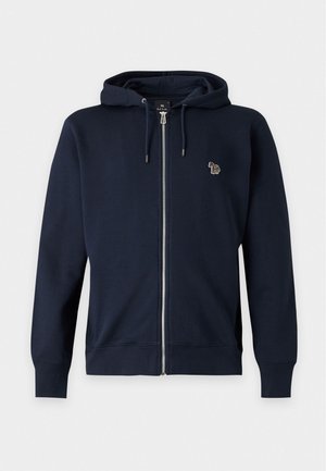 Joven de pie con las manos en los bolsillos de una sudadera azul marino con cremallera, lleva una camiseta polo beige, pantalones gris oscuro y zapatos de vestir negros sobre un fondo liso.