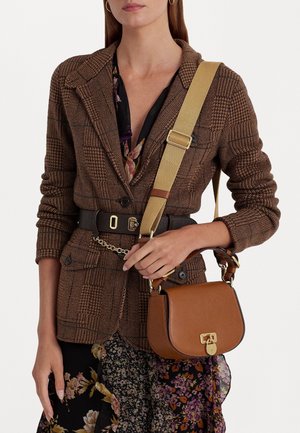 Vrouw draagt een bruin gebloemd colbert met een zwarte riem, en houdt een lichtbruine leren handtas met een brede beige schouderband.