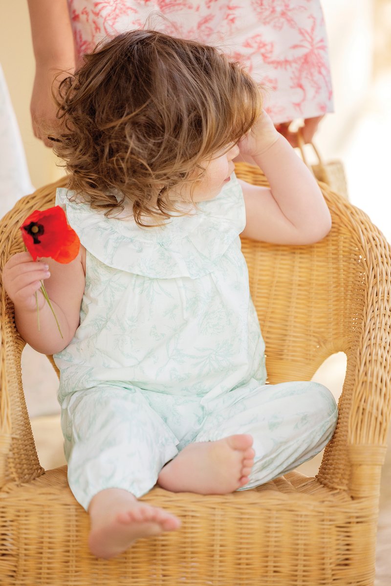 Light green floral-patterned outfit with ruffled collar, seated in a woven chair, holding a red poppy flower. Bare feet visible.