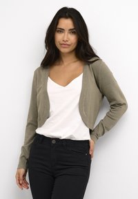 Olivgrüner Cardigan mit verkürztem Design, glatter Textur und offener Front. Über einem weißen V-Ausschnitt-Shirt getragen und mit schwarzen Jeans kombiniert.