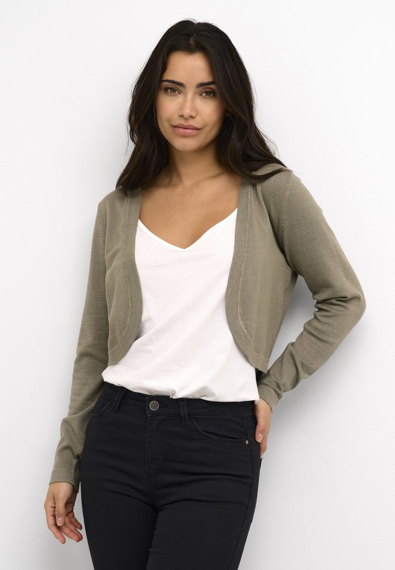 Olivgrüner Cardigan mit verkürztem Design, glatter Textur und offener Front. Über einem weißen V-Ausschnitt-Shirt getragen und mit schwarzen Jeans kombiniert.