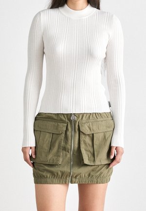 Femme portant un haut blanc à manches longues en maille côtelée et une mini-jupe vert olive avec de grandes poches avant et une fermeture éclair centrale.