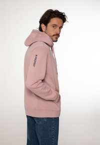 Felpa rosa con tasca frontale e cappuccio regolabile. Presenta un logo sulla manica sinistra e polsini a coste. Indossata con jeans blu.