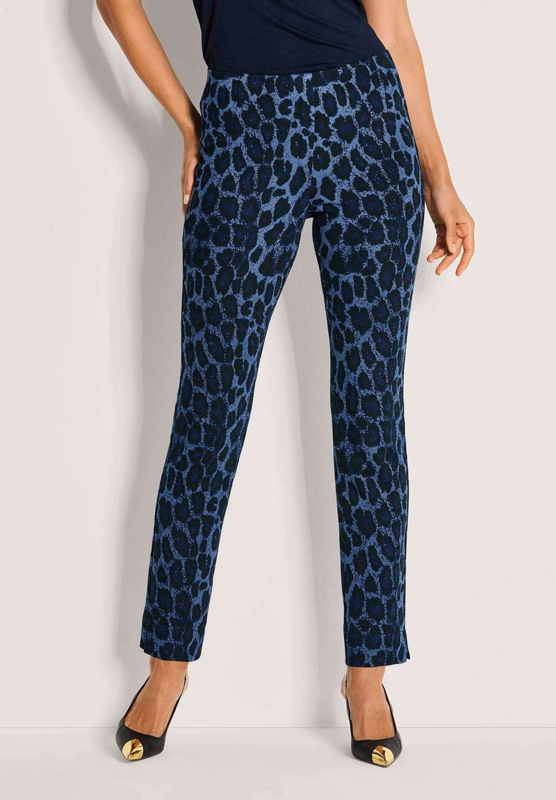 Vrouw in een aansluitende donkerblauwe panterprint broek en zwarte hakken met gouden details, staand tegen een plain beige muur.