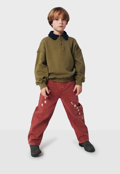 Sweatshirt vert avec col marine, trois boutons et ourlet à côtes ; pantalon rouge avec lettres blanches. Des chaussures noires complètent la tenue.