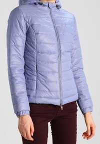 Ljuslila puffjacka med huva, som har horisontella quiltade linjer, en framställande dragkedja, elastiska ärmslut och dekorativa snördetaljer.