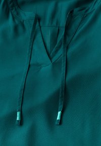 Blusa verde acqua con scollo a V, lacci regolabili e morbida trama liscia. Dettagli delle cuciture visibili attorno al colletto e all'apertura.