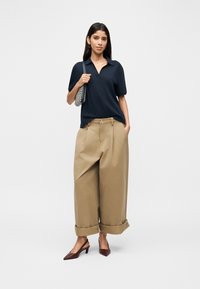 Γυναίκα με μπλε ναυτικό πόλο μπλουζάκι με κοντό μανίκι, φαρδιά μπεζ παντελόνια με μανσέτες, καφέ μυτερά τακούνια και κρατώντας μία τσάντα ώμου με σχέδιο.