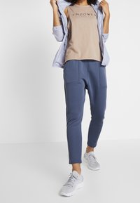Giacca azzurro chiaro sopra un top beige senza maniche con la scritta "EMPOWER", abbinata a pantaloni a gamba tapered blu scuro e scarpe sportive grigie.
