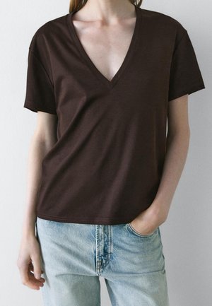 Person wearing a dark brown V-neck short-sleeve shirt and light blue jeans with one hand in pocket against a plain background.