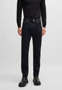 Dark blue denim jeans with a straight cut, featuring copper stitching and two front pockets. Paired with a black long-sleeve top and black boots.