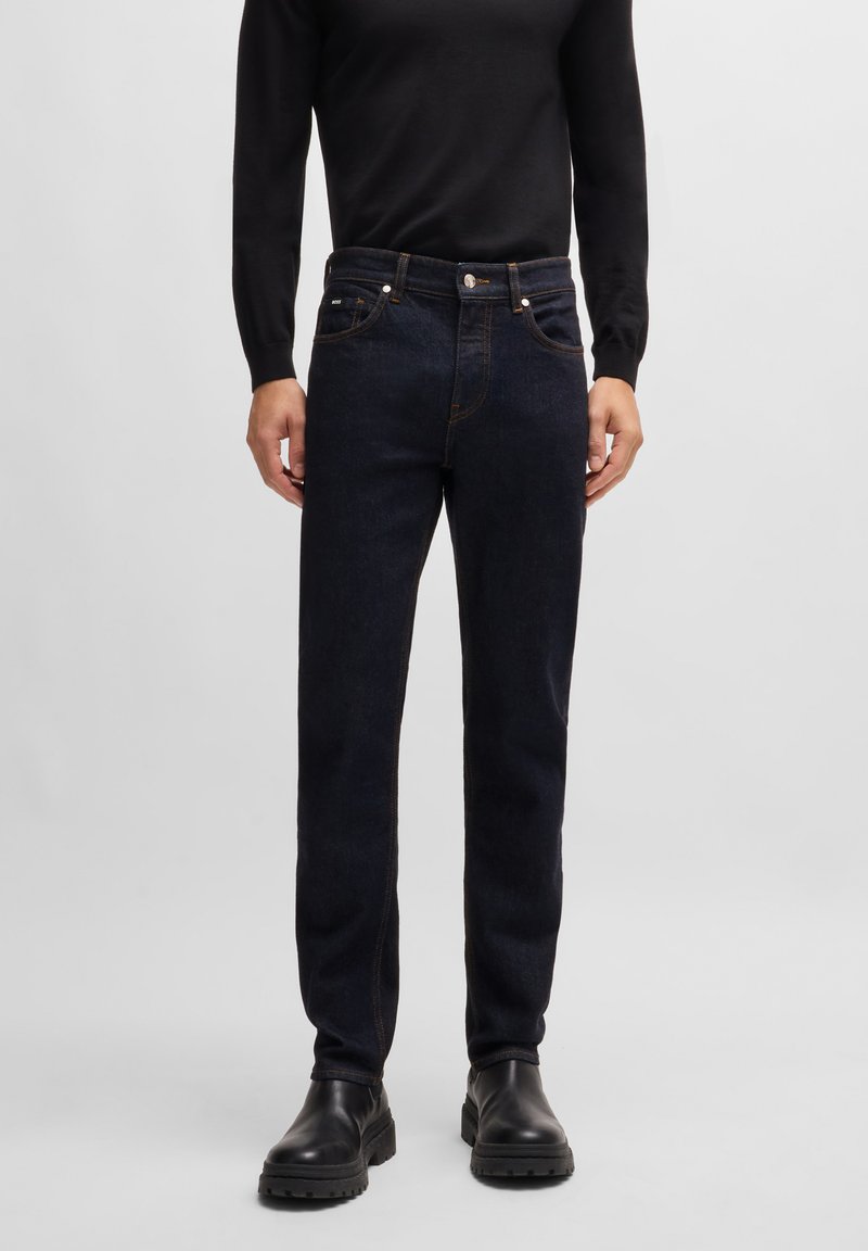 Dark blue denim jeans with a straight cut, featuring copper stitching and two front pockets. Paired with a black long-sleeve top and black boots.
