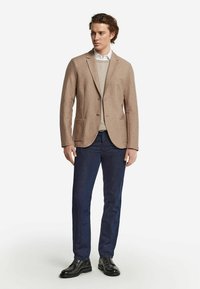 Beige kavaj med två framfickor, bärs över en ljus tröja, matchad med mörkblå jeans och svarta snörskor, stående mot en vit bakgrund.