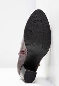 Semelle d'une botte de cheville en cuir bordeaux avec semelle en caoutchouc noir et petit talon bloc, inclinée contre un fond blanc en angle.