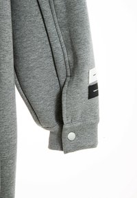 Sudadera gris con puños acanalados y botón a presión, costuras en ángulo y parches decorativos en blanco y negro.