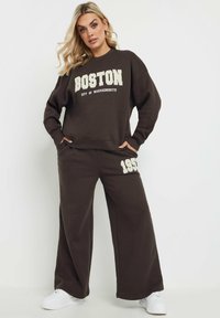 Brun sweatshirt och matchande vida sweatpants med texten "BOSTON" och "CITY OF MASSACHUSETTS", med en 1957-accent på byxorna.