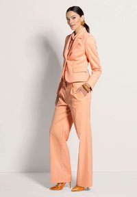 Perzikkleurig kostuum met een getailleerde blazer en een wijde broek, gecombineerd met glanzende oranje hoge hakken en grote ronde oorbellen.