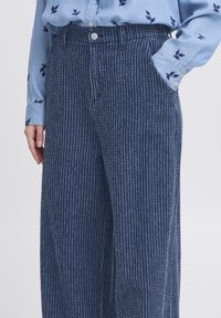 Primer plano de unos pantalones de rayas azules con textura, con botón en la parte delantera y bolsillos laterales, combinados con una camisa de manga larga floral en tono azul claro.