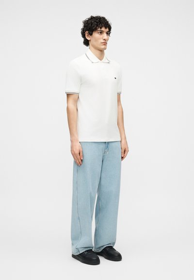 Νέος με σγουρά μαλλιά που φοράει λευκό πόλο πουκάμισο, ανοιχτό μπλε τζιν με φαρδιά πόδια και μαύρα παπούτσια, στέκεται μπροστά σε απλό φόντο.